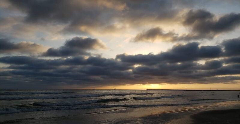 Sunset over a calm beach with gentle waves, silhouetted sailboats on the horizon, and dramatic clouds reflecting golden hues.