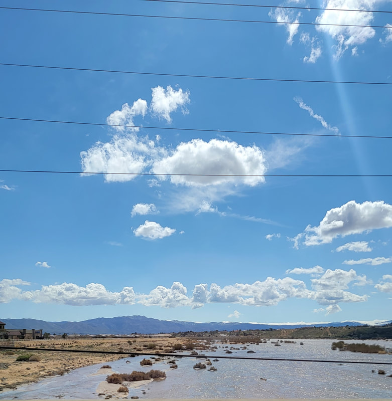 Lush, grassy landscape with a winding river under a bright blue sky, dotted with fluffy clouds and distant mountains, showcasing natural beauty.