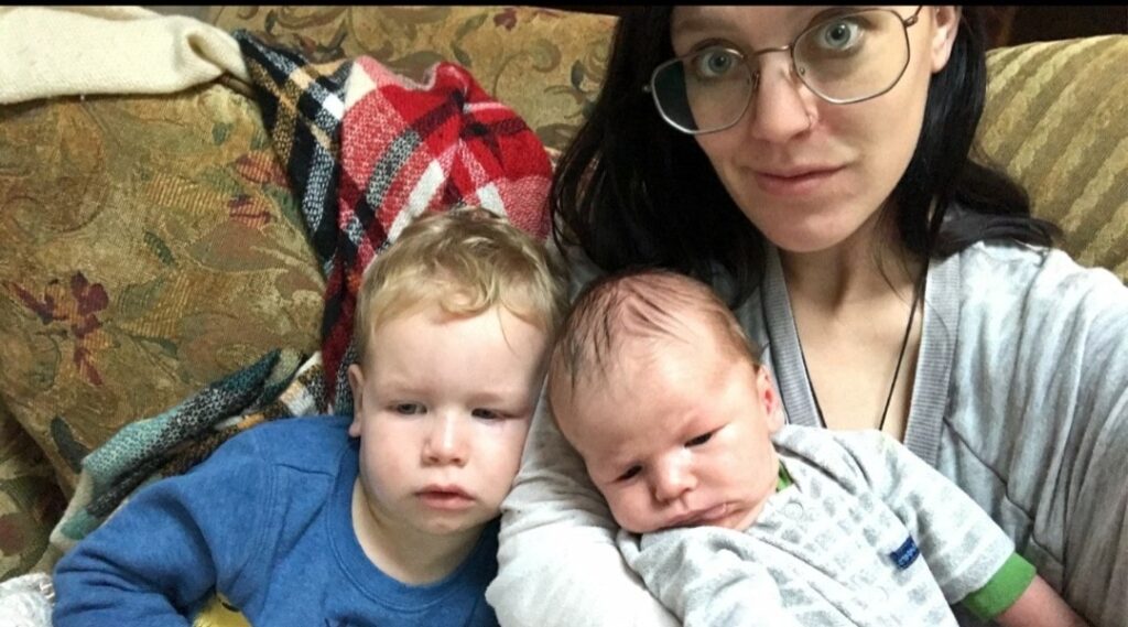 A woman sits on a couch, holding a newborn while her older child rests beside her, both looking contemplative. A cozy, patterned blanket is visible in the background.