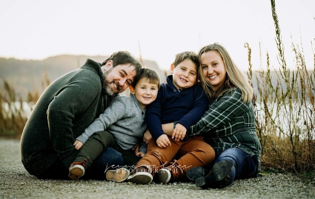 A family of four sits together on a gravel path near water, smiling warmly. The parents and two young boys are dressed casually, surrounded by tall grass and soft sunset light, capturing a moment of togetherness.