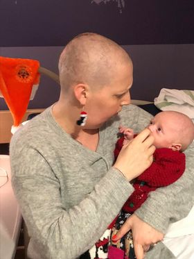A mother with a shaved head gently holds her newborn, both engaged in a tender moment. The setting is a hospital room, conveying warmth and connection.