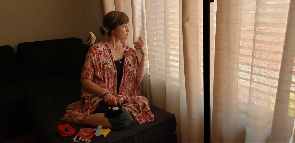 A woman in a patterned kimono sits on a couch, gazing thoughtfully out the window while a bird perches nearby. Bright sunlight filters through sheer curtains.