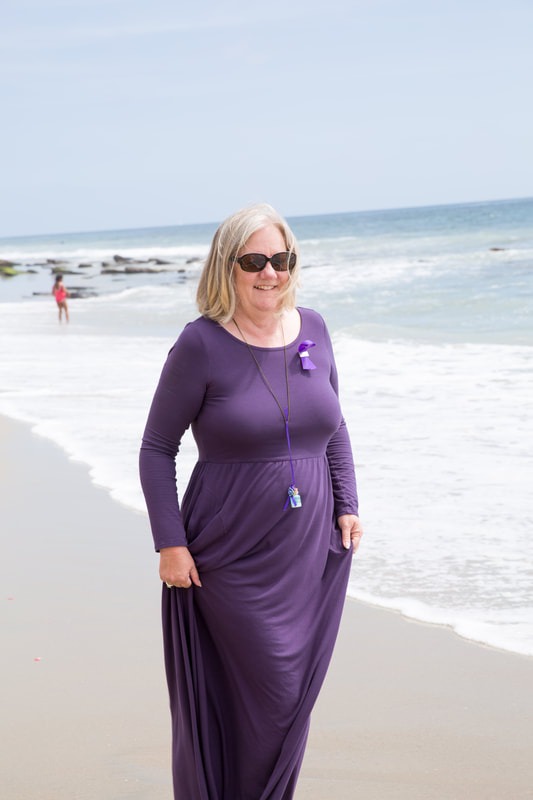 Woman in a purple dress stands on the beach, smiling confidently as waves gently lap at the shore, conveying a relaxed, joyful atmosphere.