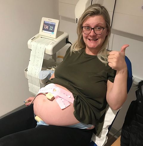Pregnant woman sitting in a medical office, giving a thumbs-up while connected to a fetal monitoring device. The setup indicates a routine checkup.