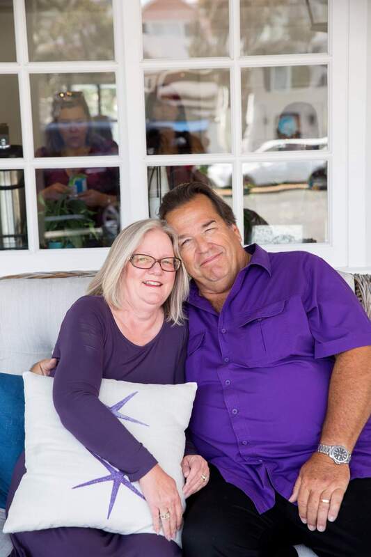 Smiling couple in matching purple outfits sit closely on a couch, holding a decorative pillow, with a bright window backdrop.