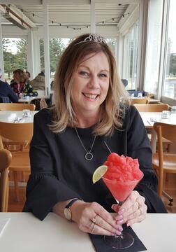 A woman in a black blouse, wearing a tiara, smiles while holding a vibrant pink slushy drink with a lime wedge, seated in a bright café.