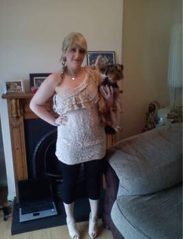 A woman with long blonde hair poses in a cozy living room, holding a small dog. She wears a light, ruffled top and black leggings, showcasing a casual yet stylish vibe.