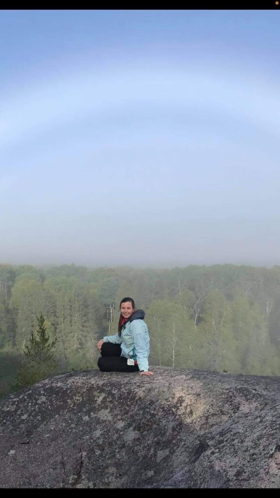 A woman sits on a large rock, smiling at the camera, with a misty forest and blue sky in the background, showcasing a serene outdoor adventure.
