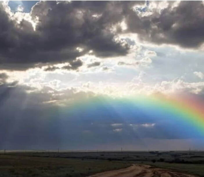 A vibrant rainbow arcs across a cloudy sky, casting colorful light over a dirt road and the surrounding landscape, symbolizing hope and beauty.