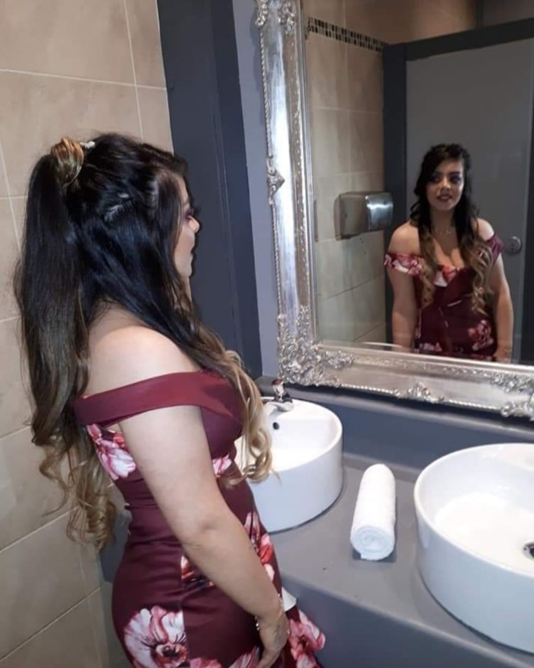A woman in a floral dress stands by a sink in a bathroom, looking at her reflection in an ornate mirror. The setting highlights her outfit and preparation.