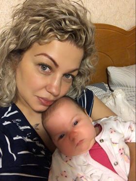 A smiling woman with curly hair holds a newborn baby on a bed. Both look at the camera, showcasing a tender moment between mother and child.