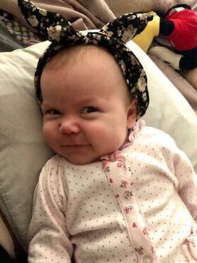 Baby wearing a floral headband with bunny ears, smiling playfully while lying on a pillow in a cozy setting.