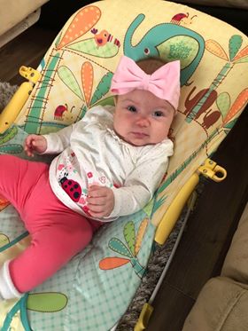 Infant girl in a colorful bouncer, wearing a white shirt and pink pants, with a large pink bow on her head, looking curiously at the camera.