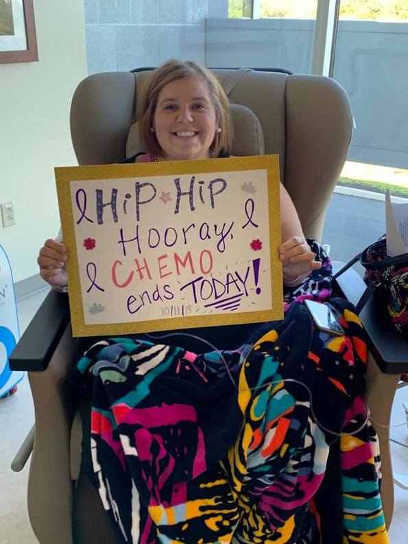 Smiling woman in a treatment chair holds a colorful sign celebrating the end of her chemotherapy. A vibrant blanket drapes over her lap.