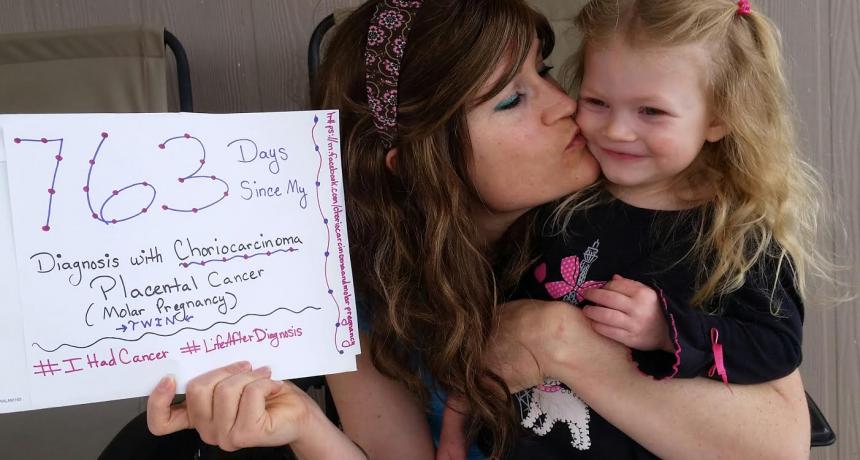 Mother kisses her daughter while holding a sign stating "763 Days Since My Diagnosis with Choriocarcinoma," reflecting on her cancer journey.